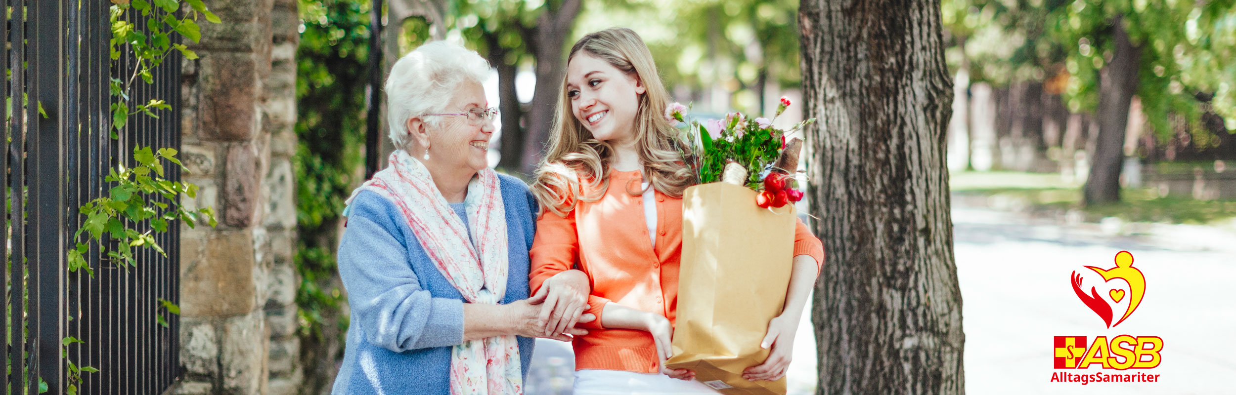 Eine junge Frau trägt eine Papiertüte mit Einkäufen. Daneben läuft eine ältere Damen, die sie anlächelt.